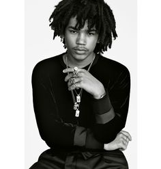 a man with dreadlocks sitting on the ground wearing a black shirt and necklace