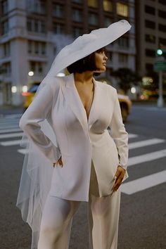 a woman in a white suit and hat standing on the street with her hands on her hips