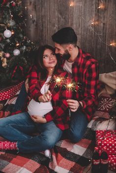 a man and woman sitting next to a christmas tree holding sparklers