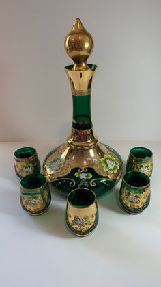 a green and gold vase sitting next to five cups on a white tablecloth covered surface