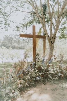 a wooden cross sitting in the middle of a field next to a tree and flowers