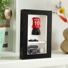 a soccer jersey and hat in a shadow box on a man's shelf next to a vase with flowers