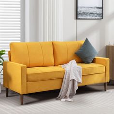 a living room with a yellow couch and white curtains on the window sill next to a potted plant