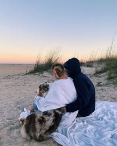 a man and woman are sitting on the beach with their dog, who is wearing a blanket