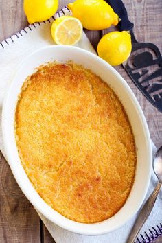 a lemon cake in a white dish on top of a wooden table next to two lemons