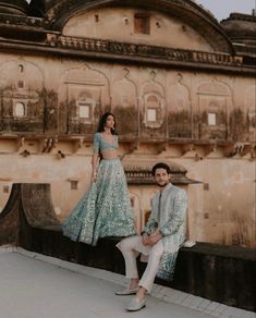 a man and woman are sitting on a ledge in front of an old building, posing for the camera