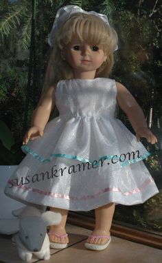 a doll with blonde hair wearing a white dress and pink shoes standing next to a stuffed animal