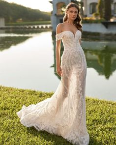 a woman in a wedding dress standing on the grass near a body of water wearing an off - shoulder gown