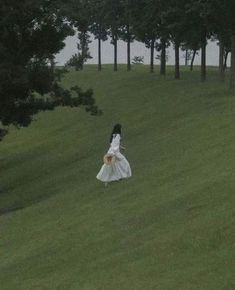 a woman in a white dress walking across a lush green field next to tall trees