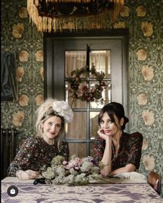 two women sitting at a table in front of a floral wallpapered room with an ornate chandelier