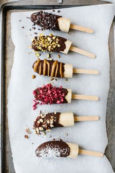 five popsicles are lined up on a sheet of wax paper with nuts and other toppings