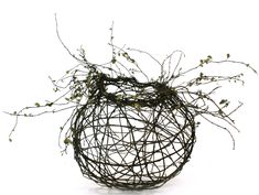 a wire vase sitting on top of a wooden table next to a white wall with plants growing out of it