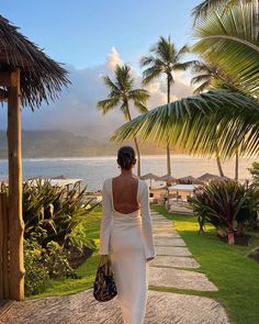 a woman in a white dress is walking towards the ocean with palm trees on either side