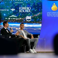 two men sitting on chairs in front of a stage talking to each other at an event