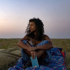 a woman sitting on top of a blanket next to a bag in the middle of a field