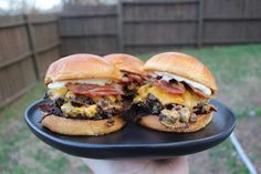 two cheeseburgers with bacon and melted cheese on a black platter in someone's hand