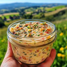 a hand holding a jar filled with food on top of a lush green field covered in yellow flowers