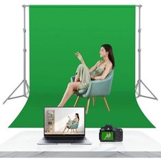 a woman sitting on a chair in front of a green screen with a laptop and camera