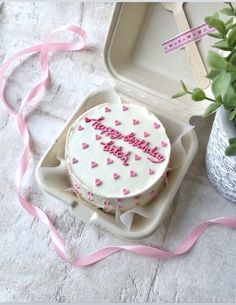 a birthday cake with pink hearts on it in a box next to a potted plant