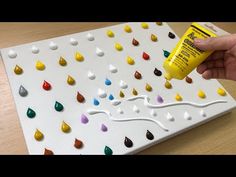 a hand holding a tube of glue next to a white board with multicolored drops on it