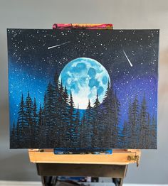 an easel with a painting on it that has a full moon and stars in the sky