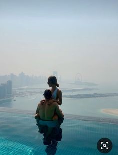 two people are sitting on the edge of a swimming pool looking out at the city