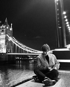 a man sitting on the ground looking at his cell phone in front of a bridge