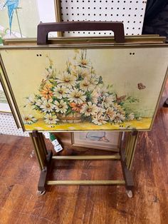 an easel with flowers painted on it sitting on a hard wood floor next to a wall