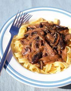 a white plate topped with pasta covered in sauce and mushrooms next to a fork on top of a blue and white striped table cloth