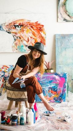 a woman sitting on top of a table next to paint and brushes in front of paintings