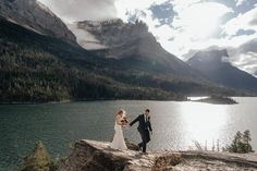 a bride and groom holding hands on the edge of a cliff overlooking a mountain lake