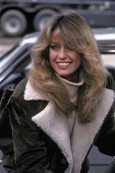 a woman with long blonde hair standing in front of a car and smiling at the camera