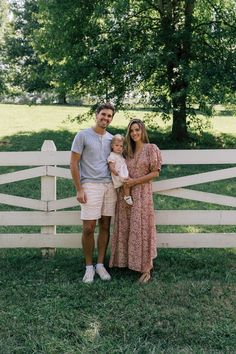 a man and woman standing next to a white fence holding a baby in their arms