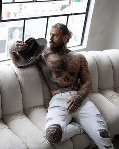 a tattooed man sitting on top of a white couch next to a black cowboy hat