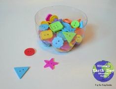 a bowl filled with lots of different colored buttons next to some shapes and sizes on a white surface