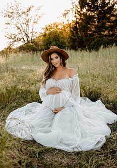 a pregnant woman wearing a white dress and hat sitting on the ground in tall grass