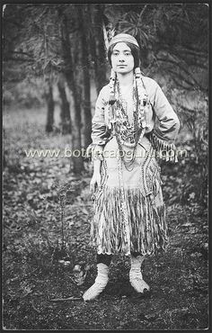 an old black and white photo of a woman in native clothing standing in the woods