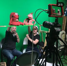 two people sitting at a table in front of a green screen with puppets on it