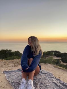 a woman sitting on top of a blanket next to the ocean with her legs crossed