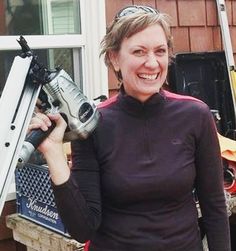 a smiling woman holding a driller in her right hand and standing next to a house