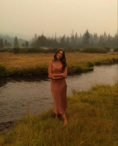 a woman standing on the side of a river next to a lush green field and forest