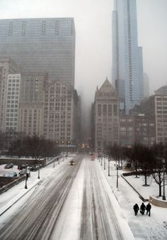 two people walking down a snow covered street in the middle of a city with tall buildings