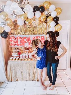 two women standing next to each other in front of a table with balloons and cake