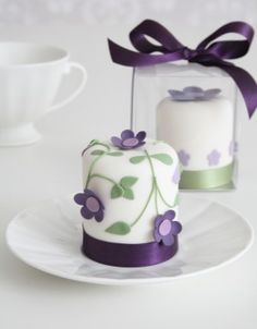 two small cakes with purple flowers on them sitting in front of a cup and saucer