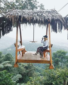 a woman sitting on top of a wooden swing in the middle of a lush green forest