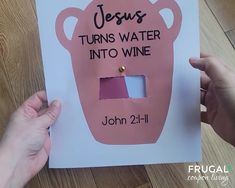 two hands holding up a pink jesus turns water into wine sign on a wooden table