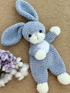 a knitted blue bunny laying next to a purple flower and cotton ball on the ground