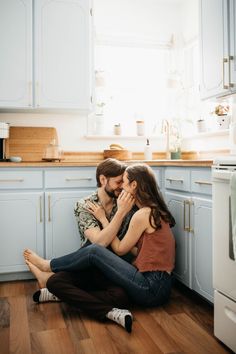 a man and woman sitting on the floor in front of a kitchen counter hugging each other