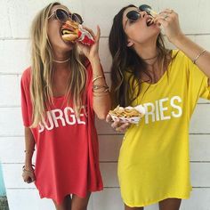 two women eating hot dogs while standing next to each other in front of a wall