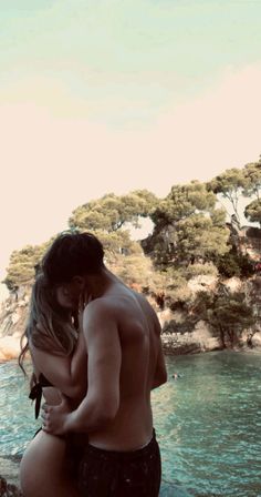 a man and woman standing next to each other near the water with trees in the background